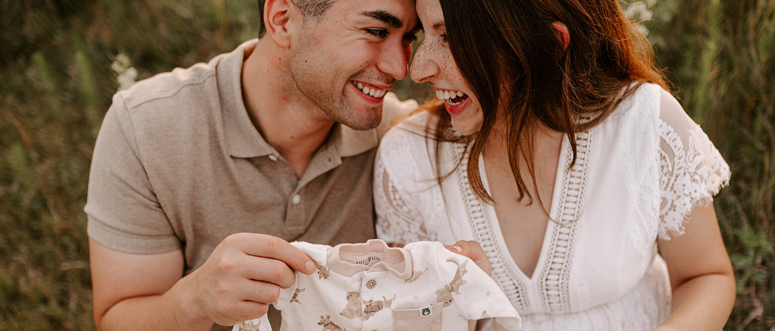Pregnant couple happily holding a baby onesie in a field